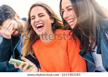 Euphoric group of friends laughing with open mouth and joking holding a smartphone in the hand sitting on an outdoors bench. Focus on the blonde haired girl.
 Royalty-Free Stock Photo #1615483585