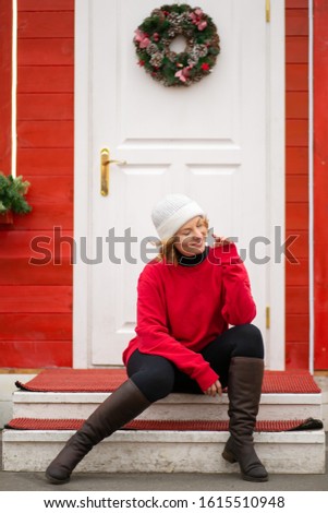 
beautiful girl in a white hat, blonde and in a red sweater, smiling, outdoors in winter, on New Year's holidays, laughing, sitting on the steps in front of a white door