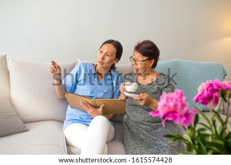 Young doctor visiting elderly woman at home. Happy grandma in glasses holding cup of coffee and watching album with photos with her nurse. Nurse and elderly women in light room