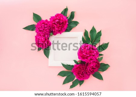 Delicate composition with wooden photo frame with white blank canvas and pink purple peony flowers with green leaves on the cold pink background. Artwork mockup with copy space