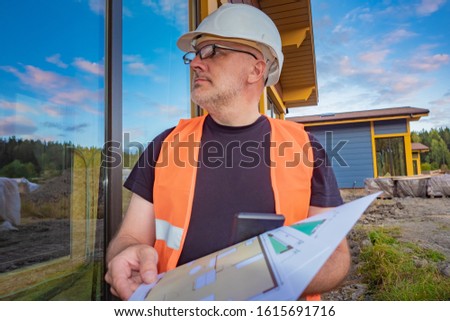 The customer evaluates the quality of construction work. The man is holding in his hands the construction documents. Construction engineer on site. A man looks at the panoramic Windows of the cottage.