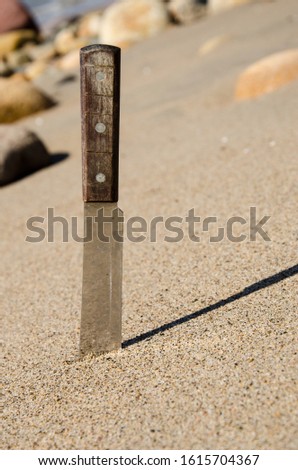 Large knife stuck in sand vertically with rocks in the background.