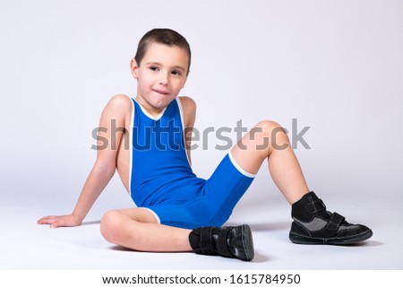 A boy athlete in sportswear and wrestling equipment sits on the floor and looks at the camera cheerfully on a white isolated background.