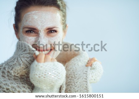 happy modern woman in roll neck sweater and cardigan with white facial mask as part of winter skin care covering in clothes isolated on winter light blue background.