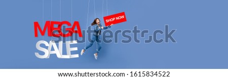 Excited beautiful Asian woman puppet on strings holding BUY NOW sign with MEGA SALE texts hanging on light blue banner background with copy space