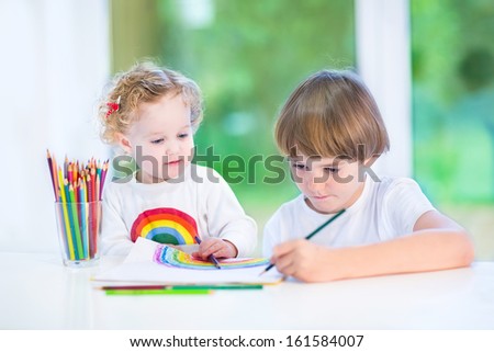 Little toddler girl watching her brother drawing with colorful pencils at a white desk next to a big window