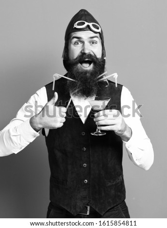 handsome bearded pilot or aviator man with long beard and mustache on happy face holding glass of alcoholic beverage in vintage suede leather waistcoat with hat, and glasses on red studio background