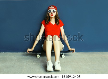 young woman sitting on skateboard wearing baseball cap, shorts on city street over blue wall background