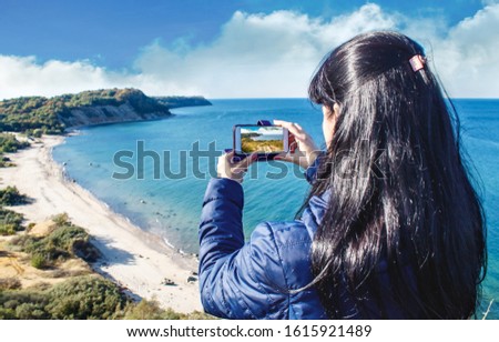 brunette woman in blue jacket photographing seascape by smartphone on sunny autumn day