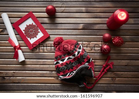Image of red snowflake in frame, decorative toy balls, cap, burning candle and rolled paper wrapped with red silk ribbon on wooden background