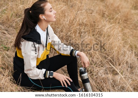 Photo of nice brunette woman in sportswear using earpods and holding water bottle while sitting on dry grass