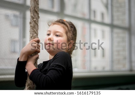 The child goes in for sports in the gym. Portrait of a schoolgirl against the background of large windows.A serious little girl is trying to climb up a thick old rope