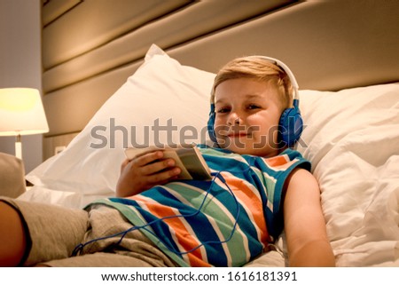 Cute boy with headphones using smart phone while lying on the bed and looking at camera. 