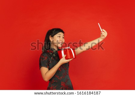 Taking selfie with gift. Happy Chinese New Year 2020. Asian young girl's portrait isolated on red background. Female model in traditional clothes looks happy. Celebration, holiday, emotions. Copyspace