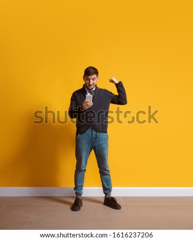 Winning game, bet. Young caucasian man using smartphone, serfing, chatting, betting. Full length portrait isolated on yellow background. Concept of modern technologies, millennials, social media.