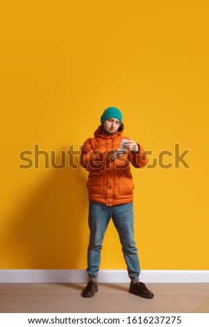 Reading news. Young caucasian man using smartphone, serfing, chatting, betting. Full length portrait isolated on yellow background. Concept of modern technologies, millennials, social media.