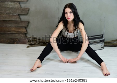 The concept of a glamorous female portrait. Portrait of a pretty brunette girl with long hair with excellent makeup in jeans and a T-shirt on a gray background sitting on the floor near the wall.