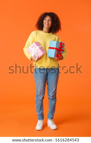 Full-length vertical shot dreamy and cute attractive african-american female looking around as going christmas, new year holiday party as secret santa, holding gifts, orange background
