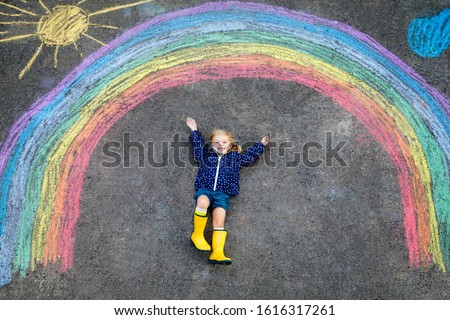 Happy little toddler girl in rubber boots with rainbow sun and clouds with rain painted with colorful chalks on ground or asphalt in summer. Cute child having fun. creative leisure