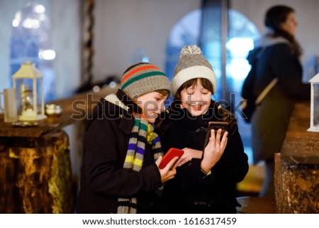 Two little kids, boy and girl having fun on traditional Christmas market during strong snowfall with taking photos with mobile smartphone or phone. Happy children, siblings and best friends