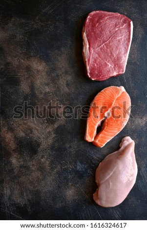 Raw marbled meat, red fish and chicken breast on a dark slate, stone or concrete background.