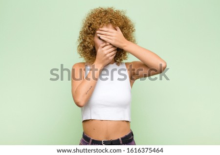 young afro woman covering face with both hands saying no to the camera! refusing pictures or forbidding photos against green wall