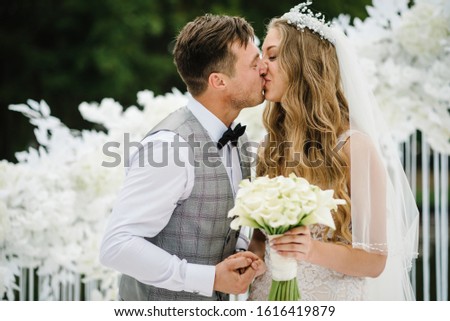 The bride and groom kissing. Newlyweds with a bouquet standing on wedding ceremony under the arch decorated with flowers and greenery of the outdoor.