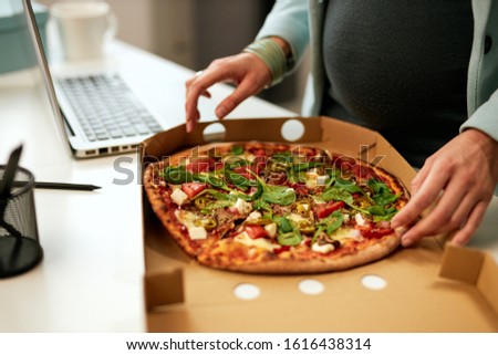 Hungry caucasian pregnant businesswoman standing in office and having pizza for lunch.