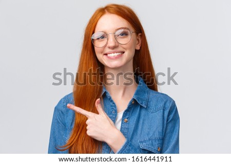 Close-up portrait cute cheerful redhead, foxy girl in glasses, showing place to click website, follow or subscribe, pointing finger left and smiling pleasant, with friendly attitude, white background
