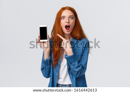 Shocked young woman saw her ex-boyfriend with someone else on internet social media. Astonished and impressed redhead woman gossiping pointing at smartphone display, drop jaw
