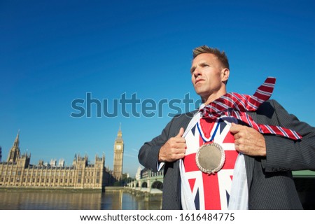 Smiling British politician pulling open his suit to reveal inner Union Jack Brexit superhero with gold medal in front of the London skyline at Westminster
