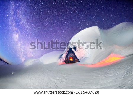 snow cornice at the top is a snow formation formed in the mountains under the influence of wind. Artistic illumination has created a fabulous fiction picture. A wooden house with a luminous window