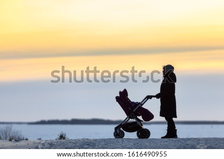 Silhouette of the woman with baby stroller standing on snowy road passing to frozen lake, winter season, sunset