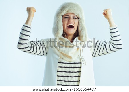 smiling stylish middle age woman in white striped sweater, scarf and ear flaps hat with raised arms rejoicing isolated on winter light blue background.