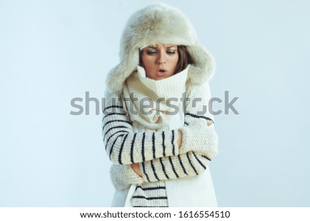 frozen stylish woman in white striped sweater, scarf and ear flaps hat against winter light blue background.