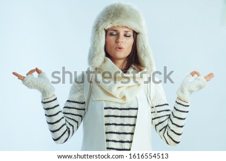 relaxed elegant 40 years old woman in white striped sweater, scarf and ear flaps hat doing yoga isolated on winter light blue background.