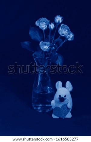 Fresh pink flowers in a glass vase and a little mouse made of textile with a pink paper heart. Painted in the classic blue color trend 2020. Selective focus.
