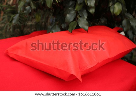 Red Color Shopping Bag Isolate on Red Color Table against natural green plant. Non Woven Lush Lava color Bag. Recyclable ECO Friendly Bag.