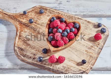 Ripe raspberries and blueberries in a wooden heart-shaped bowl on rustic cutting board. Healthy food concept. Dessert for valentines day