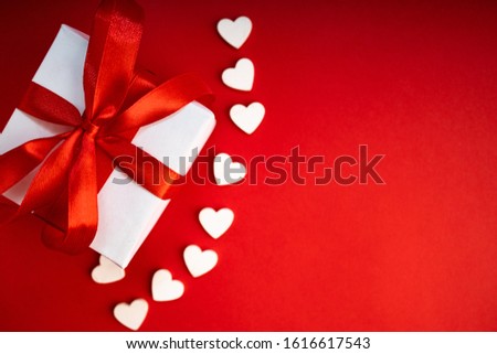 Top view of present box package decorated into the white paper and red bow-knot with signs of white wooden hearts besides. Flat lay on red background. Concept of celebrate.