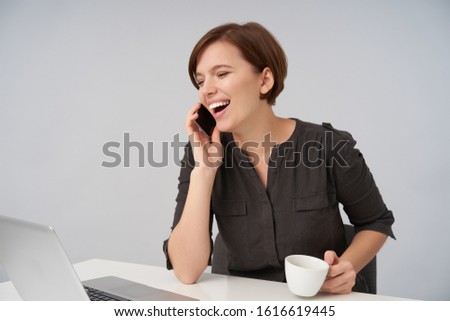 Joyful young lovely short haired brunette woman with natural makeup making break with work and calling her friend, laughing happily while sitting over white background with ceramic cup in her hand