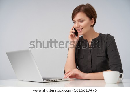 Positive young pretty short haired brunette female dressed in black shirt working at modern office with laptop and making call with her samrtphone, looking cheerfully on screen