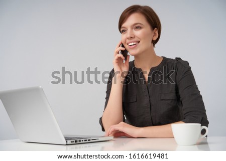 Cheerful young brown-eyed lovely brunette female with short trendy haircut smiling pleasantly while making call, sitting at table over white background with laptop and cup of coffee