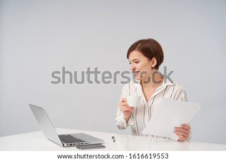 Positive young pretty brunette business woman with short trendy haircut holding cup of coffee and looking cheerfully at screen of her laptop, dressed in elegant formal clothes over white background