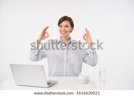 Happy young brown-eyed short haired brunette lady with casual hairstyle raising hands with ok gesture and looking positively at camera with charming smile, isolated over white background