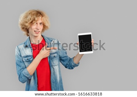 Young adult guy wearing in casual clothes looking at camera, pointing with finger on tablet computer isolated on grey background with copy space