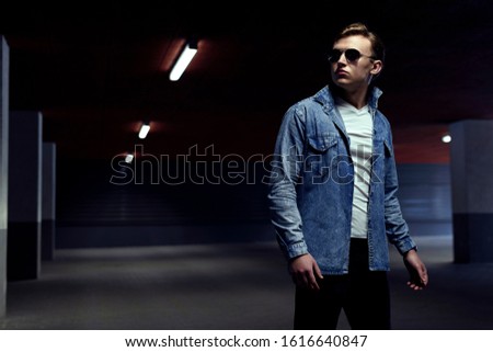 Stylish man looking somewhere. Stream of light lit his silhouette. Dressed in sunglasses, denim jacket and white shirt. Bad boy style concept. Dark background of empty underground parking.
