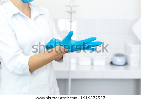 A nurse in a white coat puts on rubber gloves before a medical procedure in a bright handling room