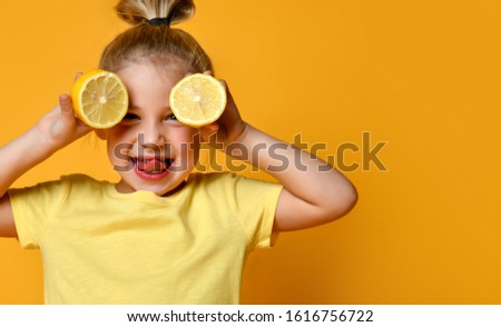 Little smiling cute blond girl in yellow t-shirt holding halves of fresh sour lemon fruit near eyes and showing tongue over yellow background. Healthy lifestyle and clean eating concept