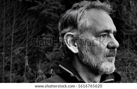 Black and white photo of a fit, attractive older Caucasian man, looking to the right, in partial profile. Short beard and gray hair. Looks calm and determined. Trees in background.